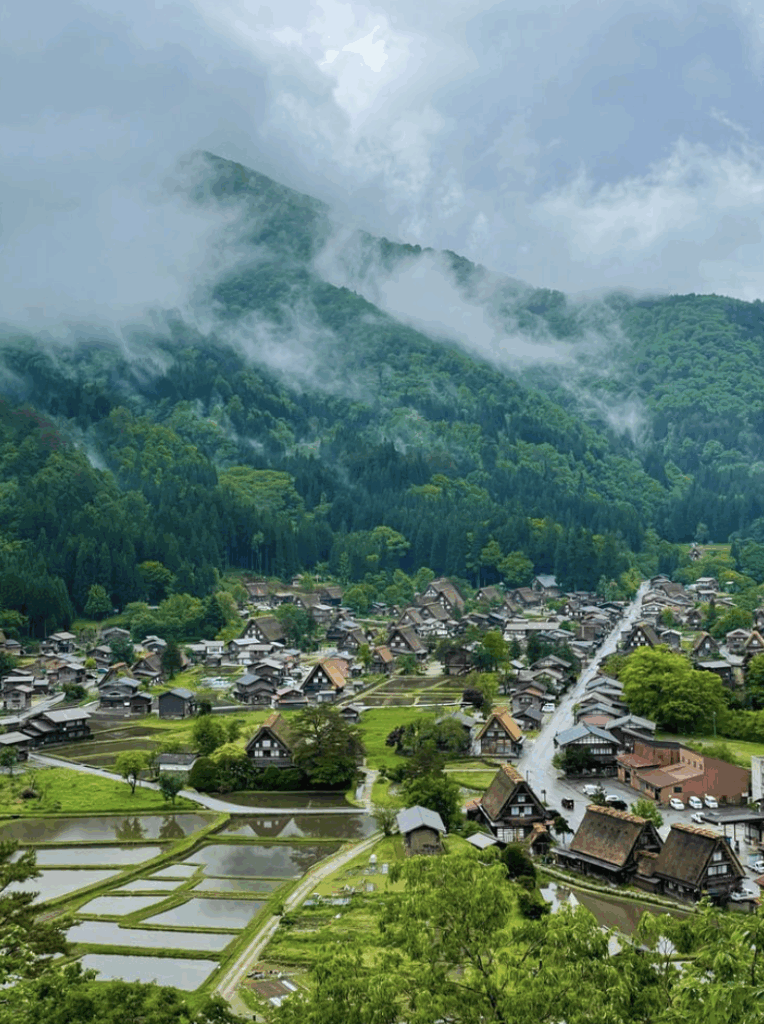 Shows a town with small hut looking houses and bright green gras and trees. Surrounding the town and in the background are mountains packed with dark green trees and clouds above and throughout the mountains.
 